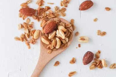 Wooden spoon filled with a mix of almonds, peanuts, and granola, placed on a white background. Scattered nuts and granola around the spoon create a natural, wholesome snack presentation.