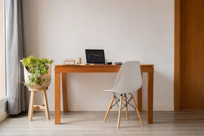 A minimalist home office with a wooden desk, white chair, and a laptop displaying a graph. A potted plant on a stool and natural light from a large window create a calm, productive workspace.