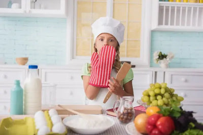 Young girl in a chef hat playfully holds a red-and-white striped oven mitt to her mouth, wooden spatula in hand, standing in a bright kitchen with eggs, flour, milk, nuts, and colorful produce on the counter.
