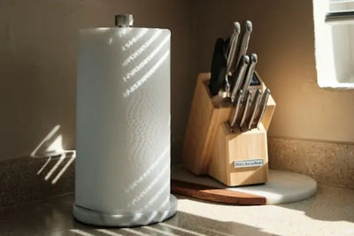 Kitchen countertop with a white paper towel roll on a marble base holder, a wooden KitchenAid knife block, and layered cutting boards. Sunlight streams through blinds, casting striped shadows on the scene.