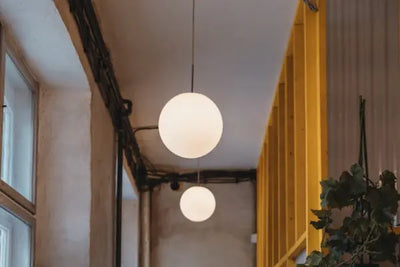 Interior with two spherical pendant lights, a large multi-pane window, concrete walls, black ceiling cables, a yellow vertical structure, and a touch of greenery from a plant, blending industrial and decorative elements.
