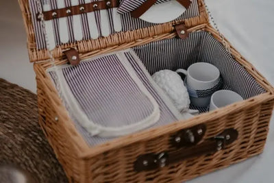 Open wicker picnic basket with purple‑white striped lining, utensils and a plate strapped in the lid, and stacked white mugs inside. Soft lighting, cozy outdoor vibe with woven mat in the corner.