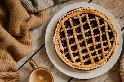 Overhead view of a lattice‑topped berry pie on a white ribbed plate, set on a rustic wooden surface with cozy knit blankets and a gold mug of coffee nearby, evoking a warm, homey vibe.