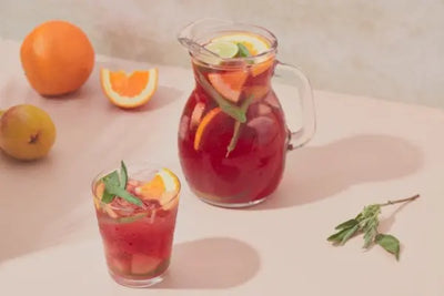 Clear glass pitcher and glass filled with red sangria, fruit slices, mint, and ice, surrounded by fresh oranges, a pear, and mint sprigs on a light surface with soft natural lighting.