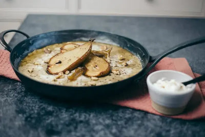 Creamy dish served in a cast iron skillet topped with sliced pears and nuts, placed on a countertop with a small bowl on the side, showcasing rustic cooking, warm flavors, and gourmet home-style presentation.