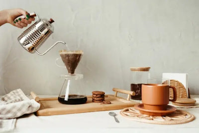 A hand pours water from a kettle into a coffee filter setup on a wooden tray with cookies and a cup. 