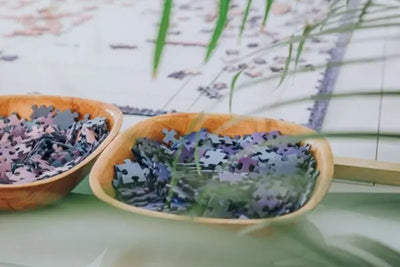 Two wooden bowls filled with purple and blue puzzle pieces on a table. A partially assembled puzzle and green leaves are blurred in the background, creating a calm, focused atmosphere.