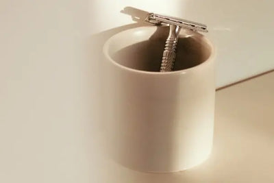 Stainless steel safety razor resting inside a ceramic mug on a bathroom counter, softly lit with warm tones, highlighting minimalist grooming setup and classic shaving tools for a clean, refined routine