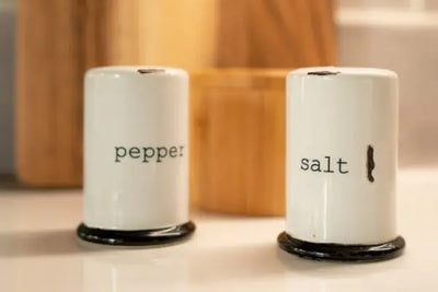 White ceramic salt and pepper shakers with black bases and text, set on a glossy surface. Blurred background features wooden kitchen items and a tiled wall, creating a clean and rustic kitchen vibe.