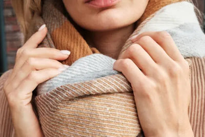 Close-up of a person adjusting a thick, knitted scarf with brown, gray, and white stripes. The scarf's cozy texture and warm colors suggest a stylish autumn or winter accessory. The blurred background keeps focus on the scarf.