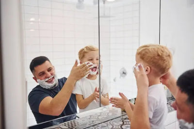 Dad and son smile in a bathroom mirror, playfully dabbing shaving cream on their faces. The father adds a bit to the boy’s cheek as the child claps, foam on his hands. White tile and a dark counter set a bright, cheerful scene of everyday family fun.