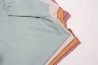 Four t-shirts that were each made in America are folded perfectly on top of each other. The colors of each t-shirt are green, beige, orange and maroon. 