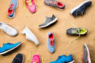 A colorful mix of athletic shoes scattered on sandy ground, featuring vibrant laces and designs in blue, pink, grey, and yellow. The casual arrangement suggests an active, outdoor lifestyle.
