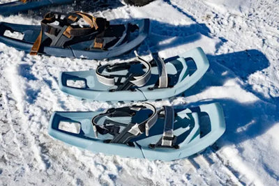 Three pairs of snowshoes on a snowy surface: two light blue pairs with black and white straps in the foreground, and a darker pair with orange and black straps behind. Featuring an open frame design, these snowshoes are ideal for winter adventures.