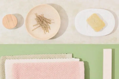 Flat lay of bathroom essentials: stacked pastel towels, a white soap dish with a cream bar, a small jar with wooden lid, a dish of bobby pins, and a slim pink container on split green/off-white background.