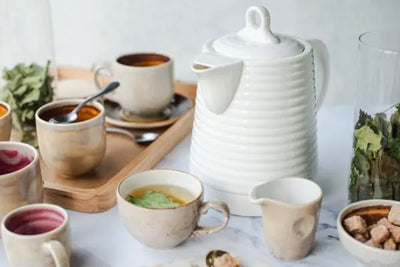 Cozy tea setup with a white ribbed teapot, assorted handcrafted ceramic cups and a small creamer, a wooden tray, a glass jar of dried herbs, and a bowl of brown sugar cubes on a light surface.