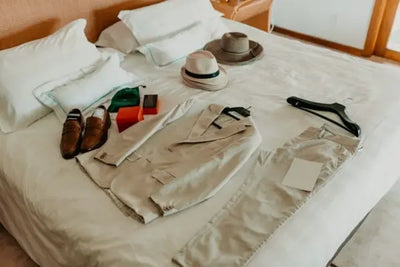Flat lay of men's formal attire on a bed: beige suit, matching trousers, brown loafers, light fedora, straw hat, and accessories. Neutral tones and soft lighting suggest preparation for a special event.