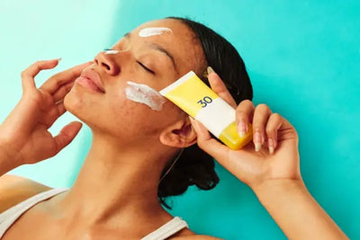 Person applying white sunscreen on their face, holding a white and yellow tube labeled SPF 30. The background is a bright aqua blue, emphasizing a fresh and vibrant skincare theme.