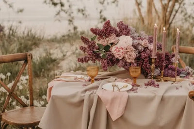 Elegant outdoor table setting with a lush purple and pink floral centerpiece, golden cutlery, amber goblets, mauve candles, and a beige tablecloth, set near water in a serene, natural setting.