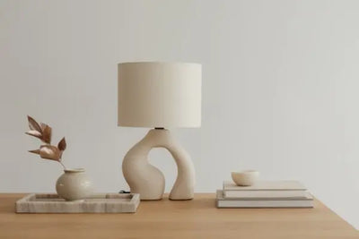 Minimalist table setup with a sculptural beige ceramic lamp, a tray holding a vase with dried reddish-brown leaves, and a stack of books topped with a small bowl. Neutral tones and soft lighting.
