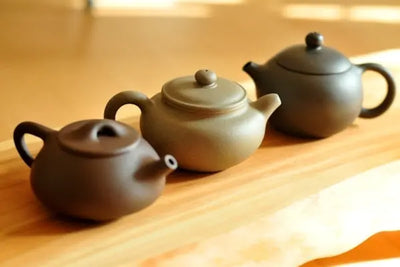 Three Yixing clay teapots in earthy tones arranged diagonally on a light wooden surface, softly lit with a shallow depth of field that highlights their rounded forms, spouts, and loop handles.