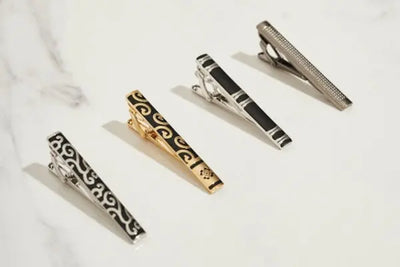 Four metallic tie clips arranged diagonally on a white marble surface. Designs include swirls, stripes, and textures in silver, gold, and gunmetal tones, showcasing elegant and versatile formal accessories.