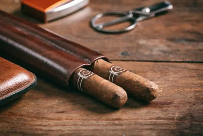 Two cigars partially in a brown leather case on a wooden table, accompanied by a metal cigar cutter and an orange-silver lighter, creating a classic and refined setup.