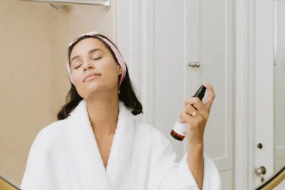 A woman in a white robe sprays her skin with American made skin toners and mists. 