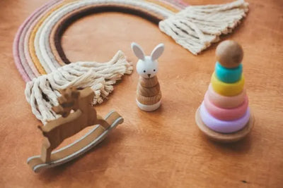 Flat lay of wooden toys on a light surface, including a rocking deer, stacking rabbit, colorful ring pyramid, and a macrame rainbow with tassels, in soft pastel and natural tones.
