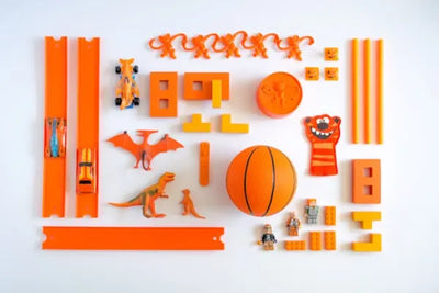 Flat lay of orange toys on a white background, including cars, a basketball, animal figures, building blocks, and a smiley face ball. The toys are neatly arranged, showcasing variety and a playful theme.