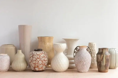 Row of assorted ceramic vases on a light wooden shelf against a plain wall—varied shapes, sizes, and textures in muted creams, tans, blush, and speckled glazes, softly lit for a curated display.