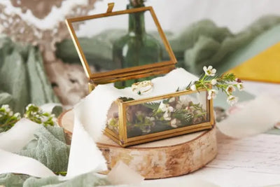 A rustic wedding-themed setup with a glass box holding white flowers, foliage, and a gold ring, placed on a wooden slab with soft fabric accents.