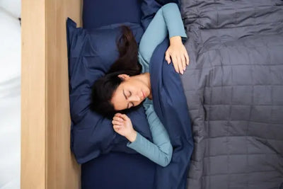 A woman peacefully sleeping under a dark blue quilted blanket, resting her head on a matching pillow. She wears a light teal long-sleeved top, creating a serene and cozy atmosphere. The image highlights comfort and relaxation.