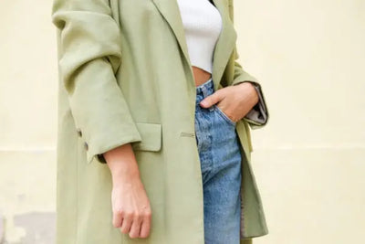 Fashion shot of a person wearing a pastel green jacket, white ribbed crop top, and high-waisted blue jeans. The outfit is casual and chic, set against a muted yellow background, highlighting the modern and stylish clothing.