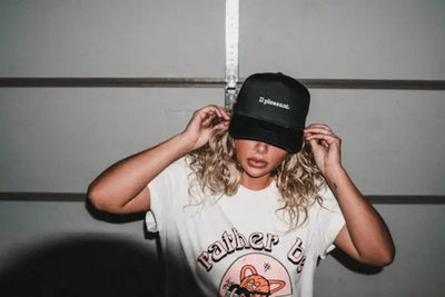 A woman sits in a garage wearing an all-black American made baseball hat. 