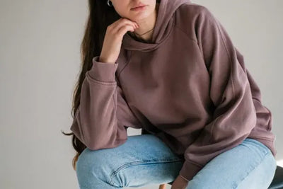 A woman sitting on a wood chair is wearing a maroon, American-made hoodie.