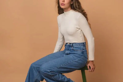 Young woman seated on a dark green stool against a beige backdrop, wearing a cream ribbed mock-neck long sleeve and high-waisted blue jeans, gazing slightly upward to her left with relaxed posture.