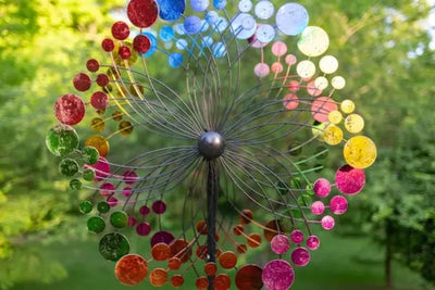 Colorful circular wind spinner with rainbow-hued discs radiating from a dark central hub. Set against a blurred green foliage background, it adds vibrant charm to an outdoor garden or yard space.