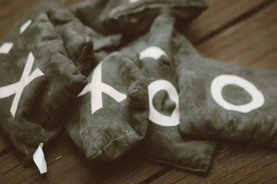 Dark gray fabric beanbags with bold white X and O symbols piled on a rustic wooden surface. Soft, diffused lighting and shallow depth of field highlight the game pieces, suggesting tic-tac-toe play.