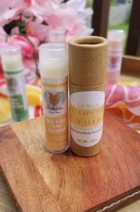 Two tubes of Bathsheba Body Works Lip Balm - My Lips Are Sealed, one in clear plastic and one in a cardboard tube, displayed on wood with a pink floral background for naturally nourishing lip care.