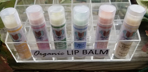 Six tubes of Bathsheba Body Works Lip Balm - My Lips Are Sealed in assorted flavors are upright in a clear acrylic holder with a front label—ideal for nourishing, natural lip care.