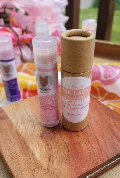 Two tubes of Bathsheba Body Works Pink Lady Lemonade Lip Balm—one in clear plastic, one in cardboard—rest on a wooden board with a floral, colorful background. Enjoy vibrant lip care made from organic and natural ingredients.