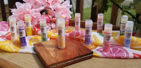 Bathsheba Body Works’ Pink Lady Lemonade Lip Balm, made with organic and natural ingredients, sits on a small wooden tray among colorful lip balm tubes, all displayed on a vibrant cloth with pink flowers in the background.