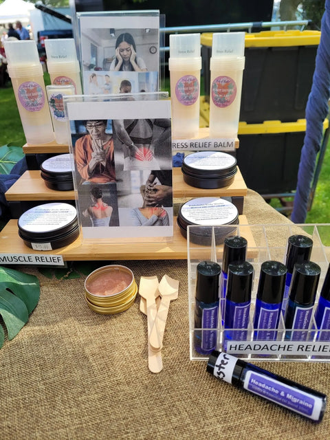 A display of Bathsheba Body Works wellness products features Moisture Balm - Muscle and Joint for stress relief, plus roll-ons for headache and migraine, all made with organic ingredients. Informational cards and wooden spatulas are included.