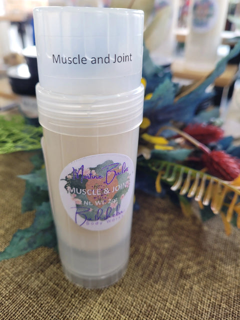 A tube of Bathsheba Body Works Moisture Balm - Muscle and Joint stands upright on a woven brown surface, offering natural pain relief, with colorful artificial foliage softly blurred in the background.