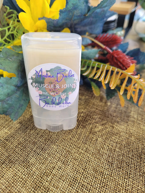 A clear tube of Moisture Balm - Muscle and Joint by Bathsheba Body Works rests on a woven surface amid artificial greenery, showcasing this natural pain relief balm made with organic ingredients.