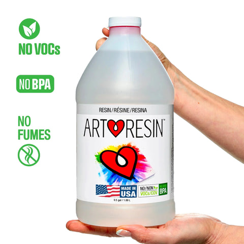 A hand holds a 1 Gal (3.78 L) ArtResin - Epoxy Resin bottle with a heart logo. The label notes: No VOCs, No BPA, No Fumes, Made in USA, and UV protection. This non-toxic kit by ArtResin is ideal for creative projects.