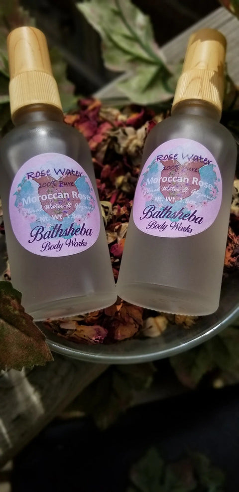 Two frosted glass bottles labeled Bathsheba Body Works Hydrosol - Rose Water sit in a metal bowl filled with dried rose petals and surrounded by leaves, emphasizing the natural essence of organic skin care.