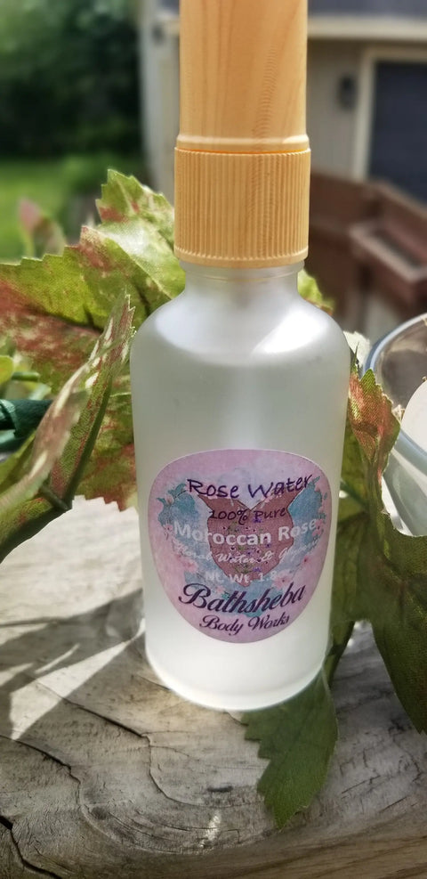 A frosted glass spray bottle of Bathsheba Body Works Hydrosol - Rose Water sits on a wooden surface among green leaves, highlighting luxurious organic skin care, with a softly blurred outdoor background.