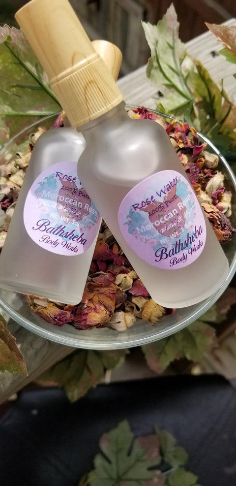 Two Bathsheba Body Works Hydrosol - Rose Water glass spray bottles rest on a bowl of dried rose petals, surrounded by green leaves—an elegant organic skin care display.
